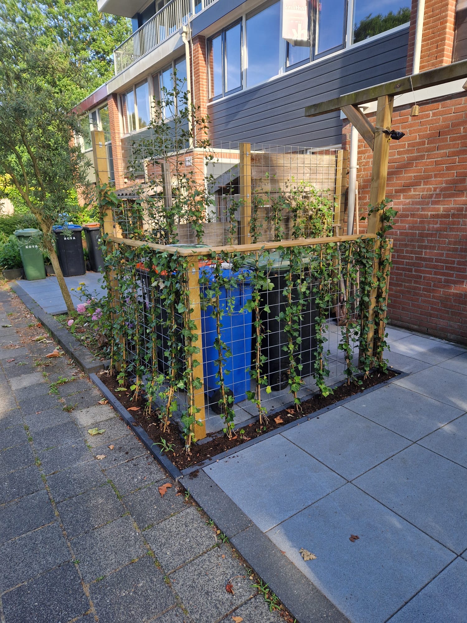 Afvalcontainers achter houten hekwerk met klimplanten